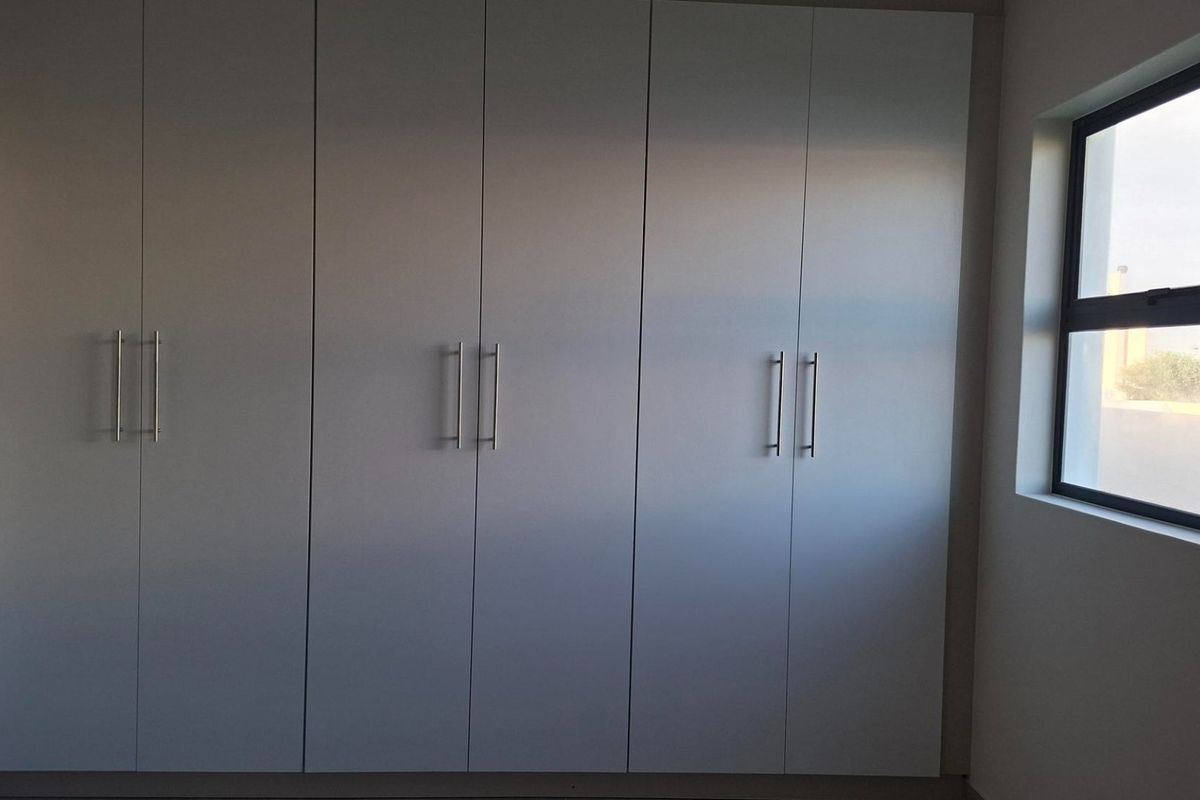 A clean, minimalist bedroom featuring floor-to-ceiling built-in cupboards with modern handles and a side window providing natural light.