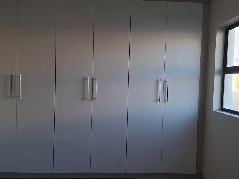 A clean, minimalist bedroom featuring floor-to-ceiling built-in cupboards with modern handles and a side window providing natural light.