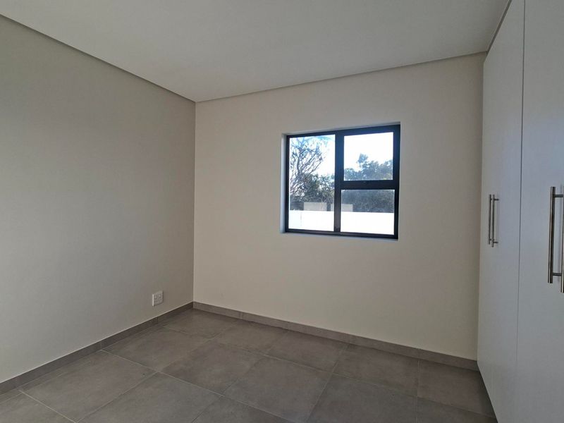 A clean, modern, and empty bedroom featuring neutral tiled flooring, built-in cupboards, and a window providing natural light.