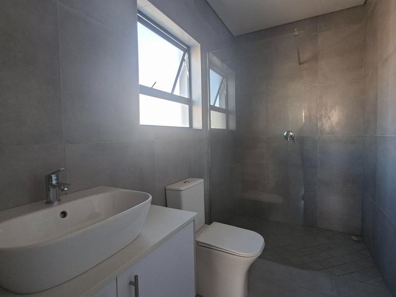 Modern bathroom featuring grey wall and floor tiles, a vessel sink with vanity cabinet, toilet, and a glass-enclosed shower.