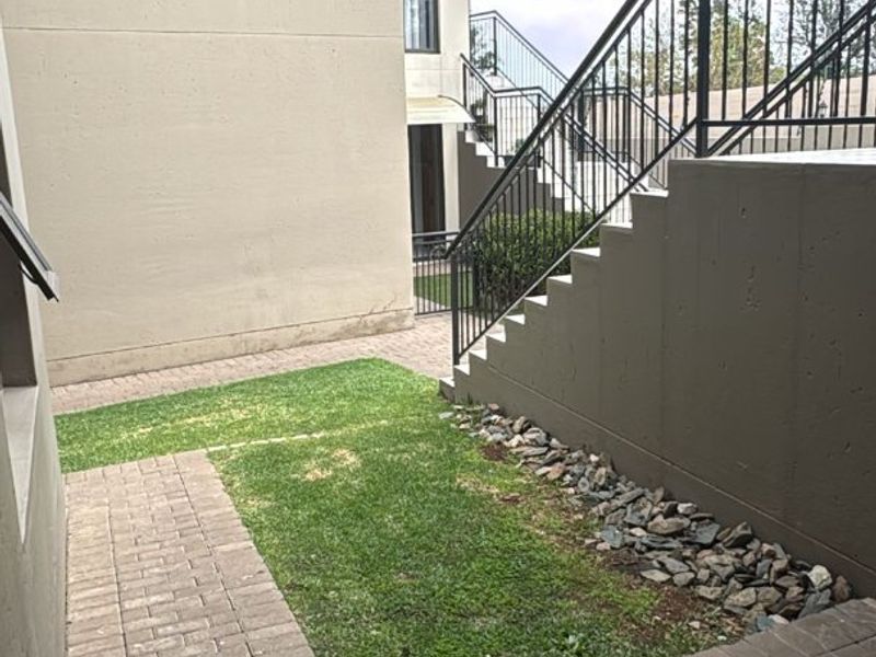 A rear exterior view of an apartment complex featuring a paved walkway, small lawn area, and an external metal staircase with railings.