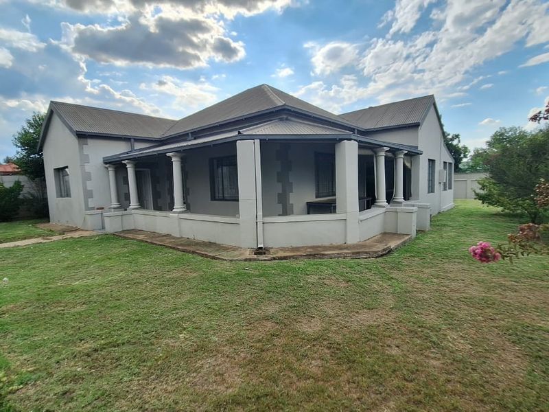 Rear view of the house featuring a wrap-around covered veranda with concrete columns, set within a spacious garden with a surrounding boundary wall.