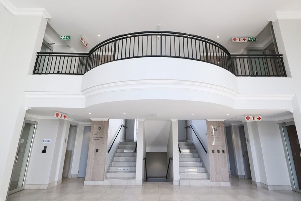 A bright, modern commercial building entrance featuring a double-volume lobby, central staircase with metal railings, and clean tiled finishes.
