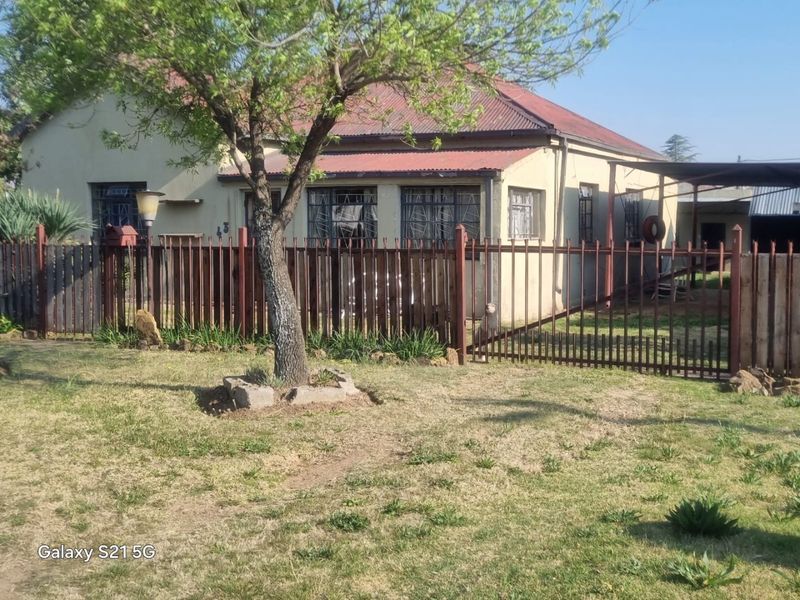 Front view of a single-story house featuring a corrugated metal roof, metal security bars on windows, and a fenced front yard with a carport.