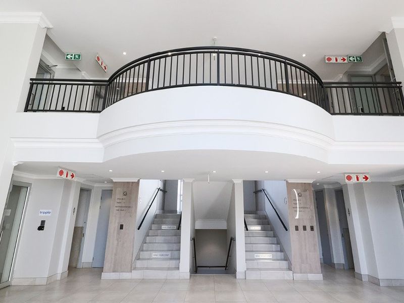 A bright, modern commercial building entrance featuring a double-volume lobby, central staircase with metal railings, and clean tiled finishes.