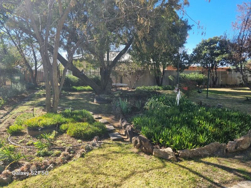 A spacious backyard garden featuring mature trees, established garden beds with stone edging, and a lawn area enclosed by metal perimeter fencing.