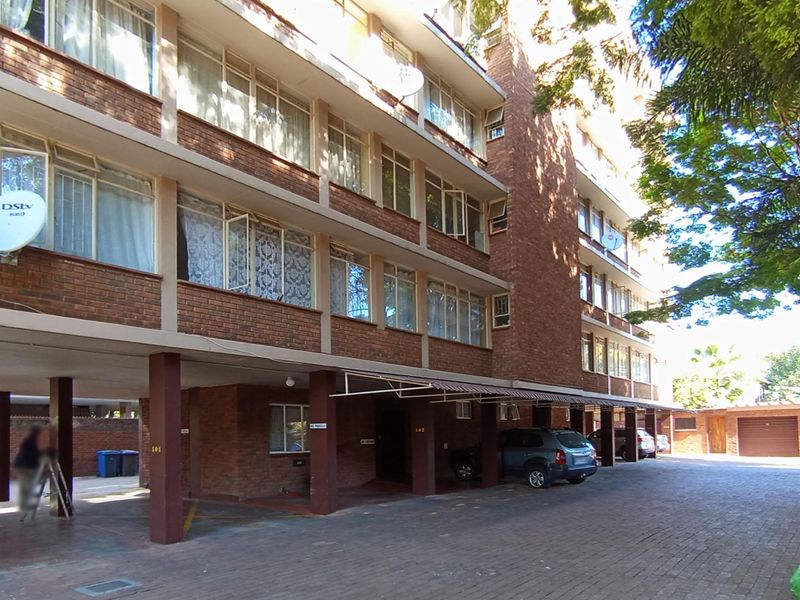 Exterior view of a multi-story brick apartment building featuring a covered ground-level parking area and paved driveway access.