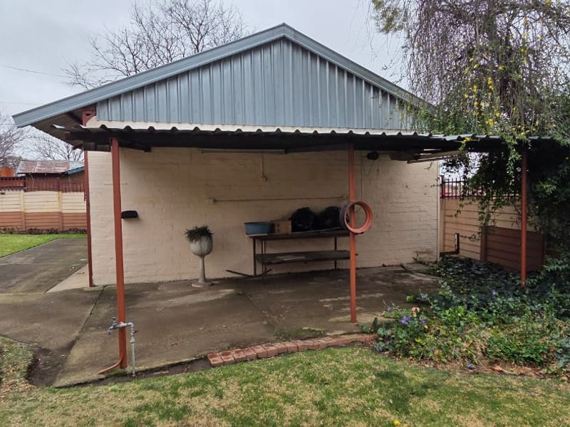 A rear exterior view of a utility structure featuring a corrugated metal roof, concrete patio area, and metal support posts in a garden setting.