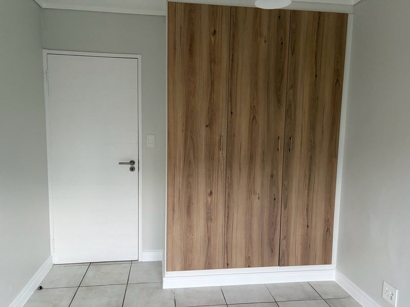 A clean, empty bedroom featuring modern wood-finish built-in cupboards, neutral wall paint, and light-colored floor tiles.