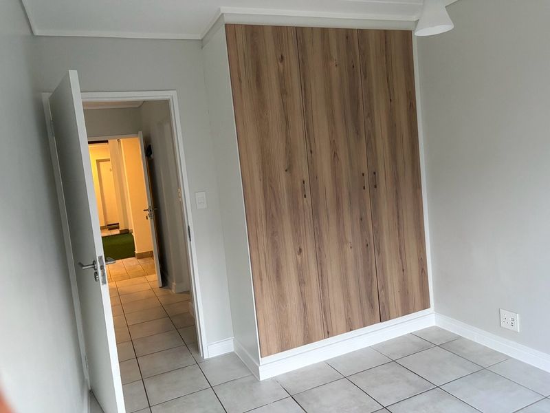 A clean, empty bedroom featuring modern wood-finish built-in cupboards, neutral tiled flooring, and a doorway leading to a hallway.