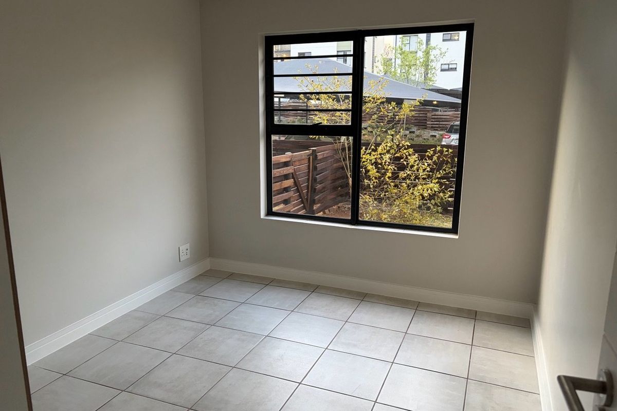 A bright, empty bedroom featuring neutral tiled flooring, clean walls, and a large black aluminium window frame overlooking the exterior.