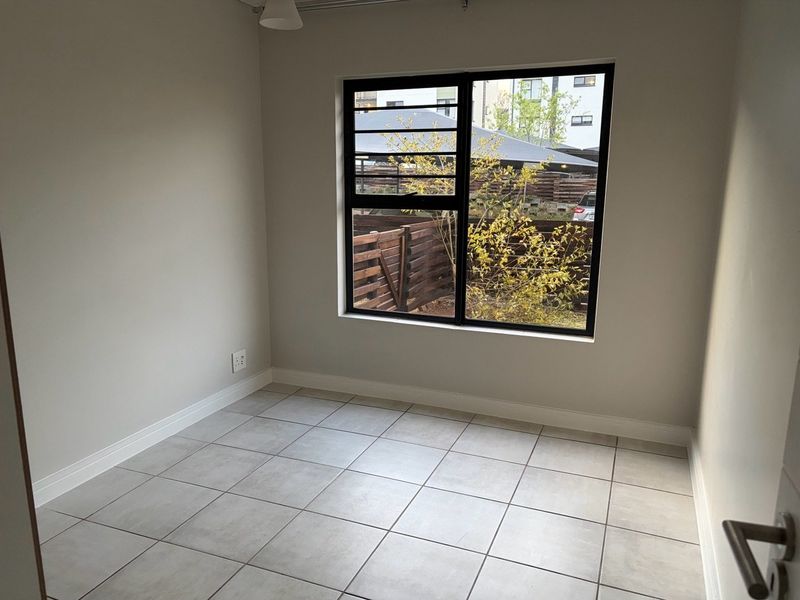 A bright, empty bedroom featuring neutral tiled flooring, clean walls, and a large black aluminium window frame overlooking the exterior.