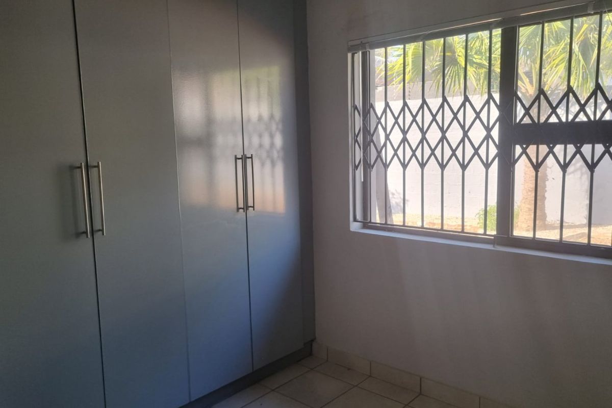 A clean, empty bedroom featuring built-in cupboards, tiled flooring, and a window fitted with a security gate.
