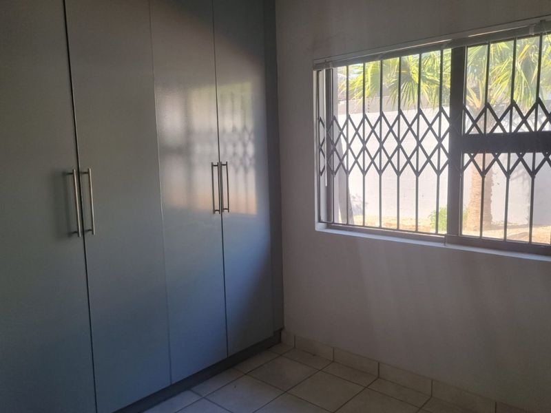 A clean, empty bedroom featuring built-in cupboards, tiled flooring, and a window fitted with a security gate.