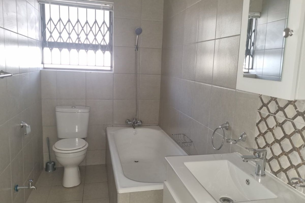 A functional bathroom featuring a bathtub, toilet, and vanity basin with neutral wall tiling and a security gate on the window.
