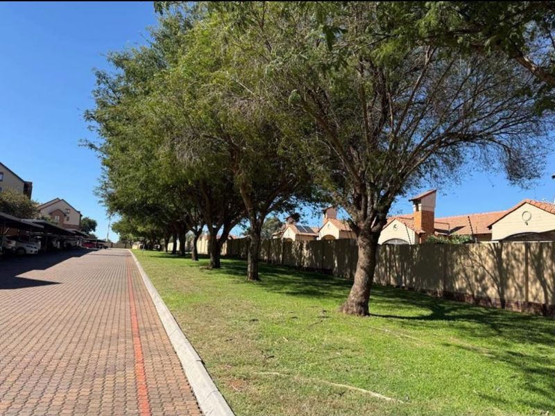 A well-maintained residential complex exterior featuring a paved driveway, covered carport parking, and a landscaped green verge with mature trees.