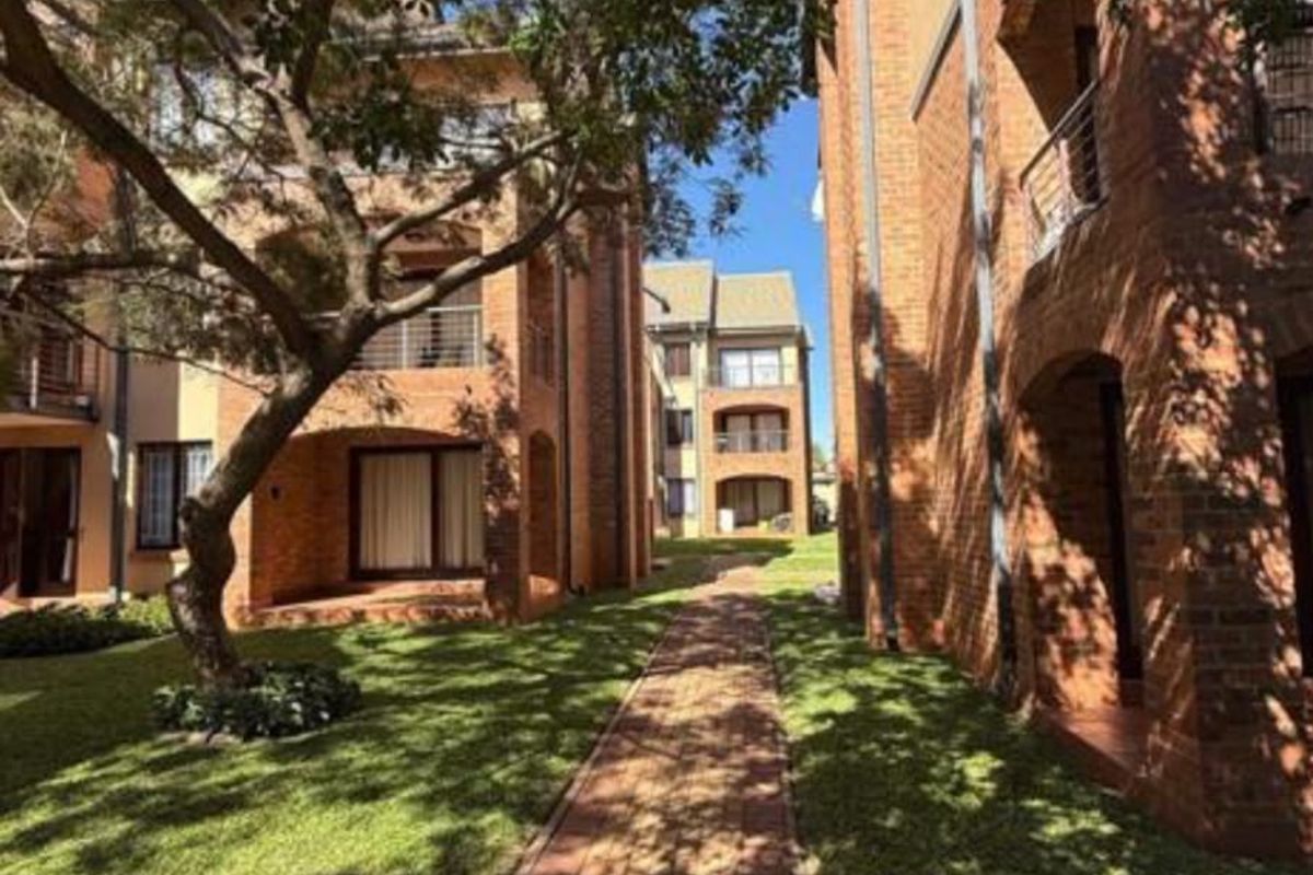 A well-maintained apartment complex exterior featuring red brick facades, private balconies, and a landscaped garden with a paved walkway.
