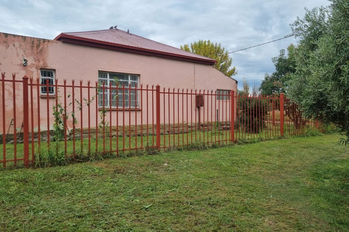 Rear view of a single-story house featuring a corrugated metal roof, security-barred windows, and perimeter metal fencing set within a garden area.