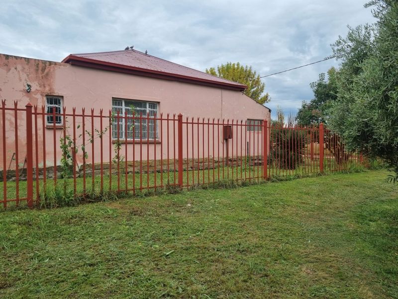 Rear view of a single-story house featuring a corrugated metal roof, security-barred windows, and perimeter metal fencing set within a garden area.