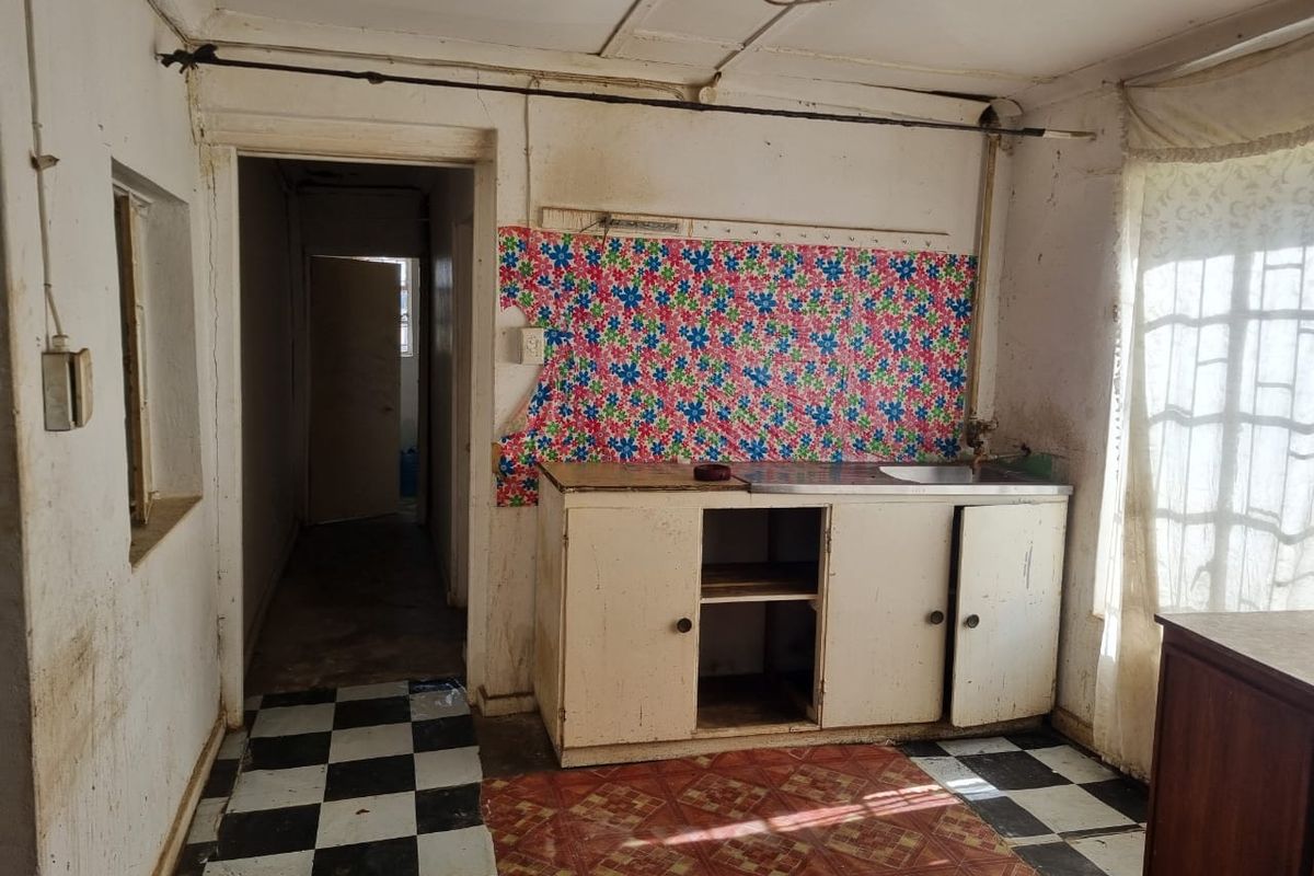 A dilapidated kitchen space featuring basic cabinetry, a stainless steel sink, and worn flooring, requiring significant renovation and repair to restore functionality.
