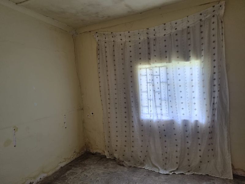 An empty bedroom featuring a window with security bars, showing significant signs of wear including wall cracks and damp staining.