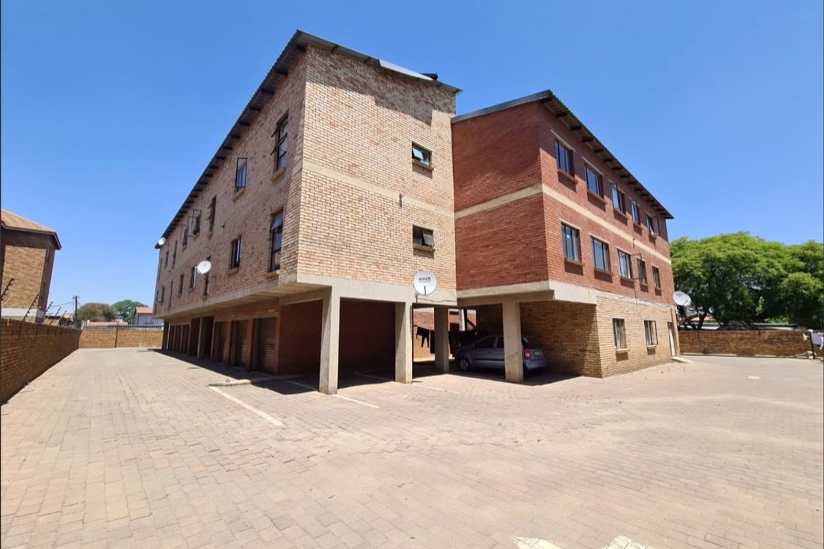 Exterior view of a multi-story face brick apartment building featuring covered ground-level parking bays and a paved driveway area.