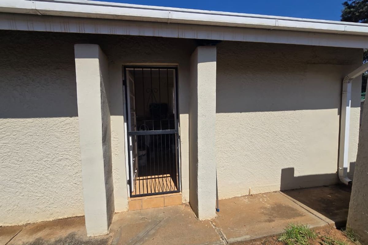 A rear exterior view showing a concrete patio area with a security gate entrance and plastered wall finish.