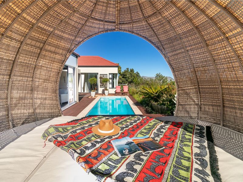 A view from a shaded wicker cabana overlooking a private swimming pool and timber deck area in a well-maintained garden setting.