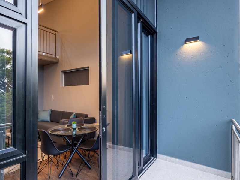 A modern balcony featuring sleek sliding glass doors, contemporary wall lighting, and a metal safety railing, overlooking a lush green exterior.