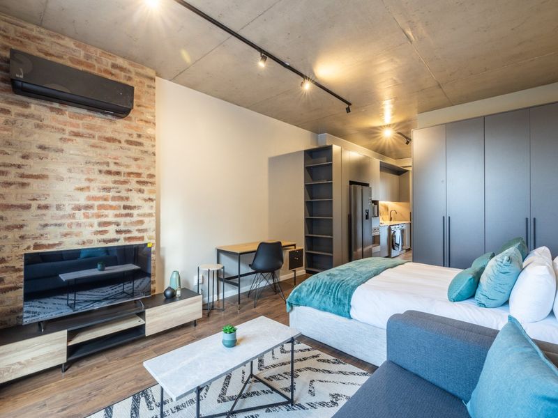 Modern open-plan studio apartment featuring an exposed concrete ceiling, brick feature wall, track lighting, and integrated cabinetry with a compact kitchen area.