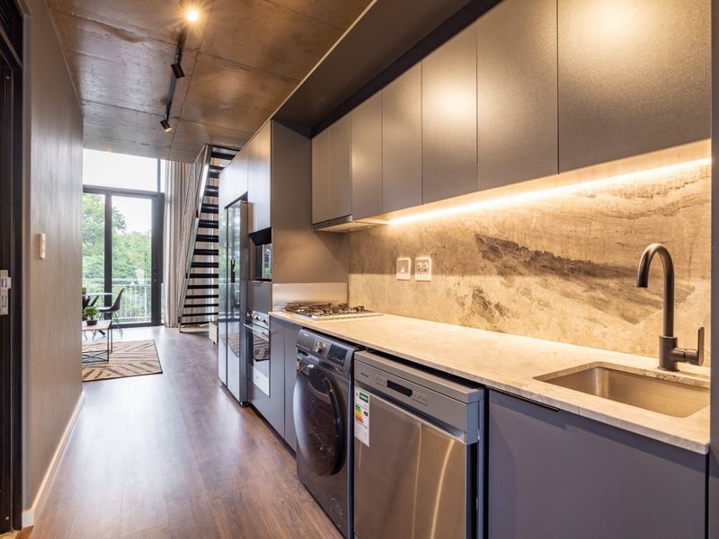Modern open-plan kitchen featuring sleek grey cabinetry, stone finishes, integrated appliances, and an exposed concrete ceiling, seamlessly connecting to the living area.