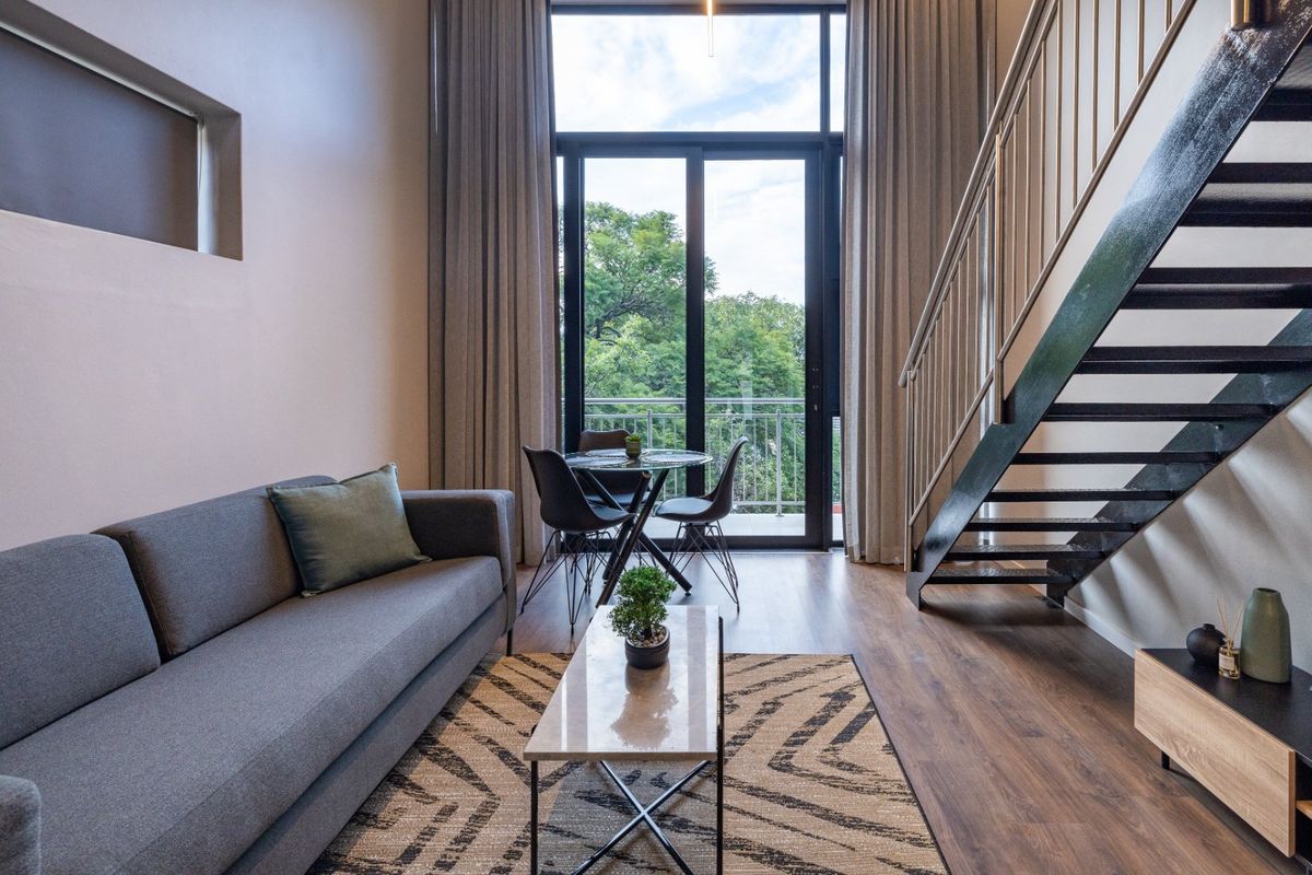 Modern double-volume living area featuring laminate flooring, a sleek metal staircase, and large sliding glass doors leading to a private balcony.