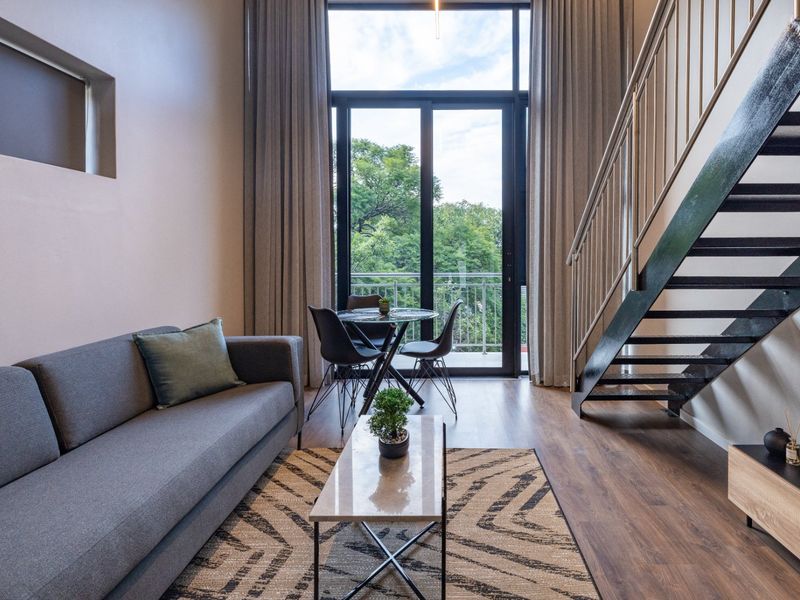 Modern double-volume living area featuring laminate flooring, a sleek metal staircase, and large sliding glass doors leading to a private balcony.