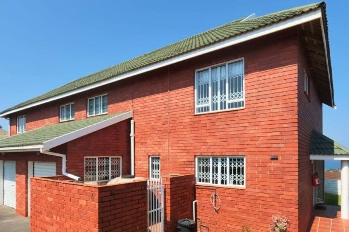 Rear view of a double-story face brick townhouse featuring a tiled roof, security bars on windows, and a paved patio area.