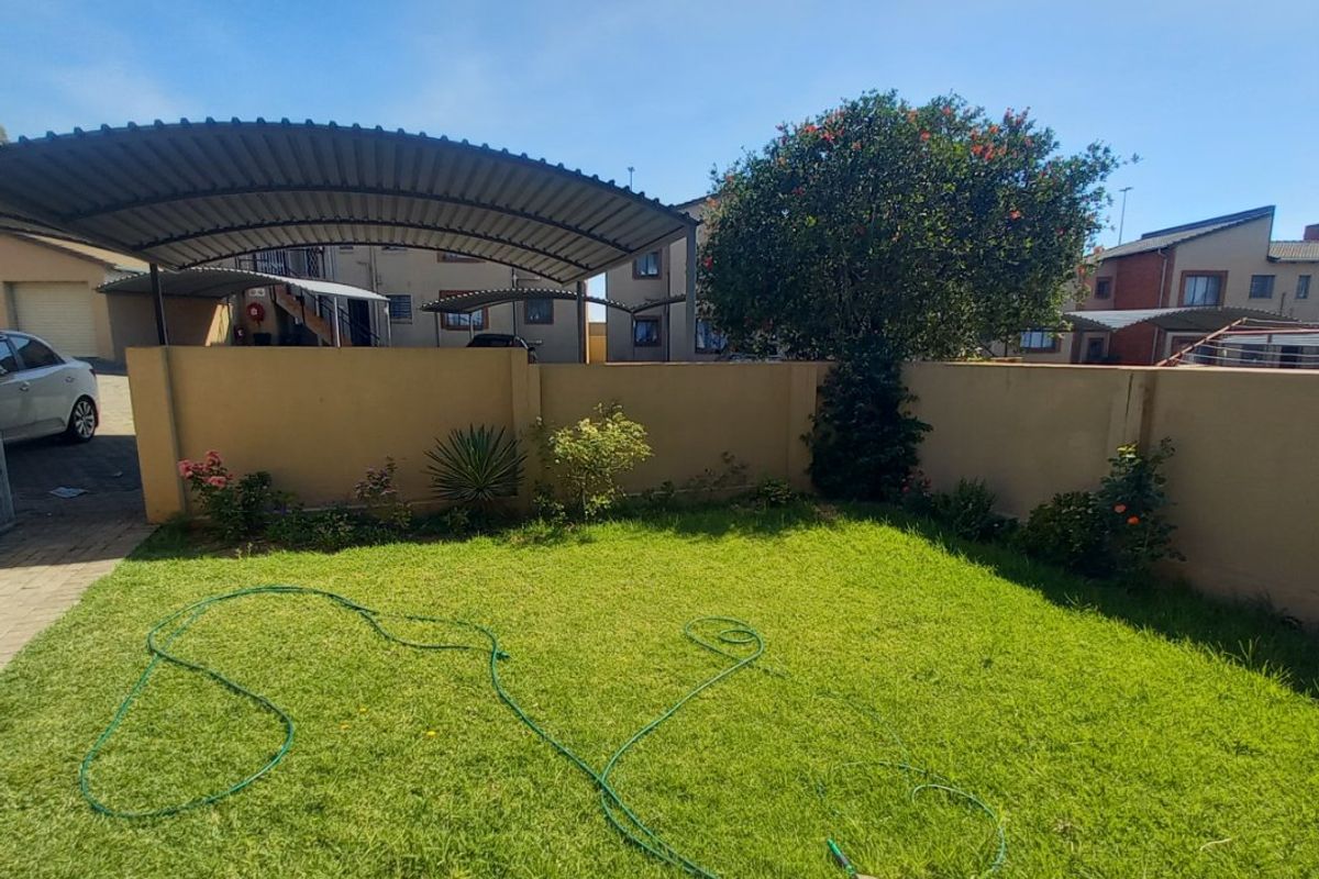 A private rear garden area featuring a lawn, paved section, and a covered carport structure enclosed by a boundary wall.