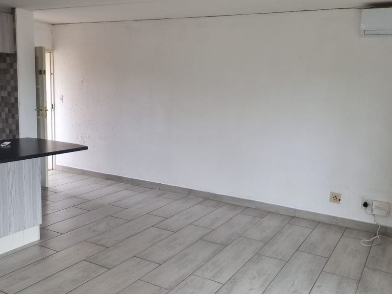 An open-plan living area featuring light-toned tiled flooring, a compact kitchen with a breakfast bar, and a wall-mounted air conditioning unit.