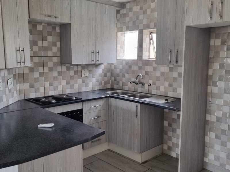 A functional kitchen featuring light wood-grain cabinetry, dark laminate countertops, a built-in electric hob and oven, and a tiled backsplash.
