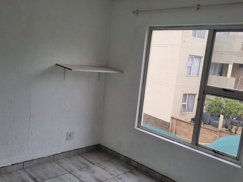 An empty bedroom featuring tiled flooring, a wall-mounted shelf, and an aluminium window frame with views of the surrounding apartment complex.