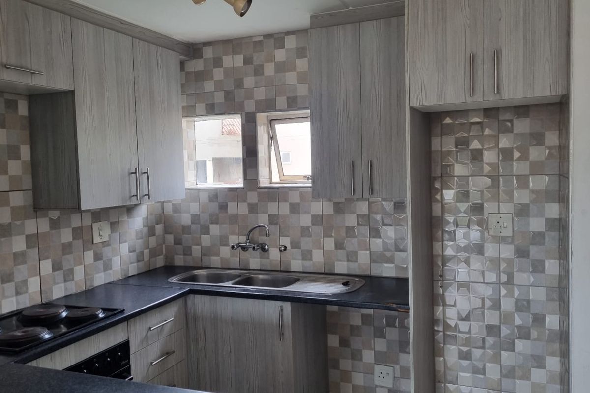 A functional kitchen featuring light wood-grain cabinetry, dark laminate countertops, a double sink, and a built-in electric hob with a decorative tiled splashback.