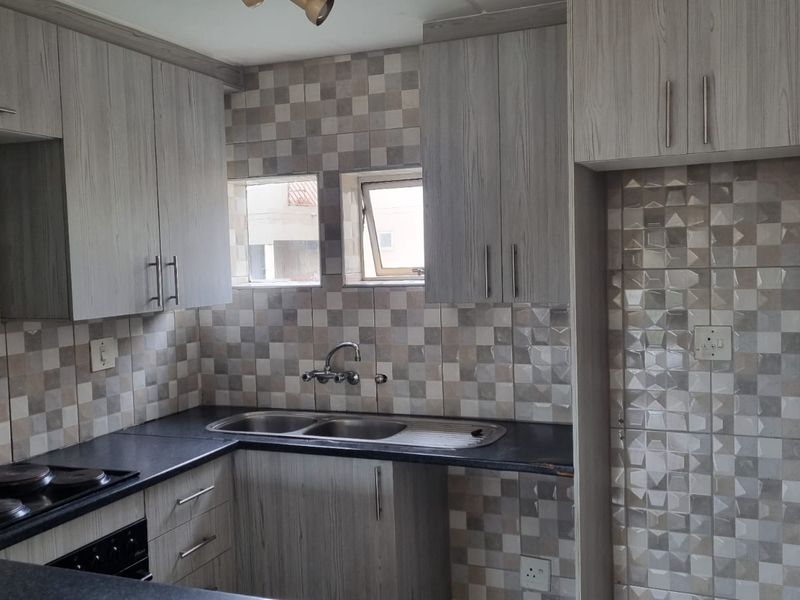 A functional kitchen featuring light wood-grain cabinetry, dark laminate countertops, a double sink, and a built-in electric hob with a decorative tiled splashback.