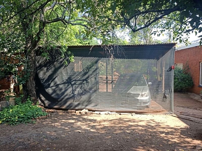 A simple outdoor carport structure featuring a corrugated metal roof and shade cloth sides, situated on a dirt surface adjacent to a brick building.