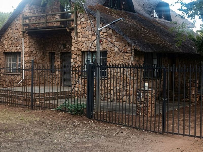 A rustic stone-clad farmhouse featuring a traditional thatched roof, a front balcony, and secure metal perimeter fencing.