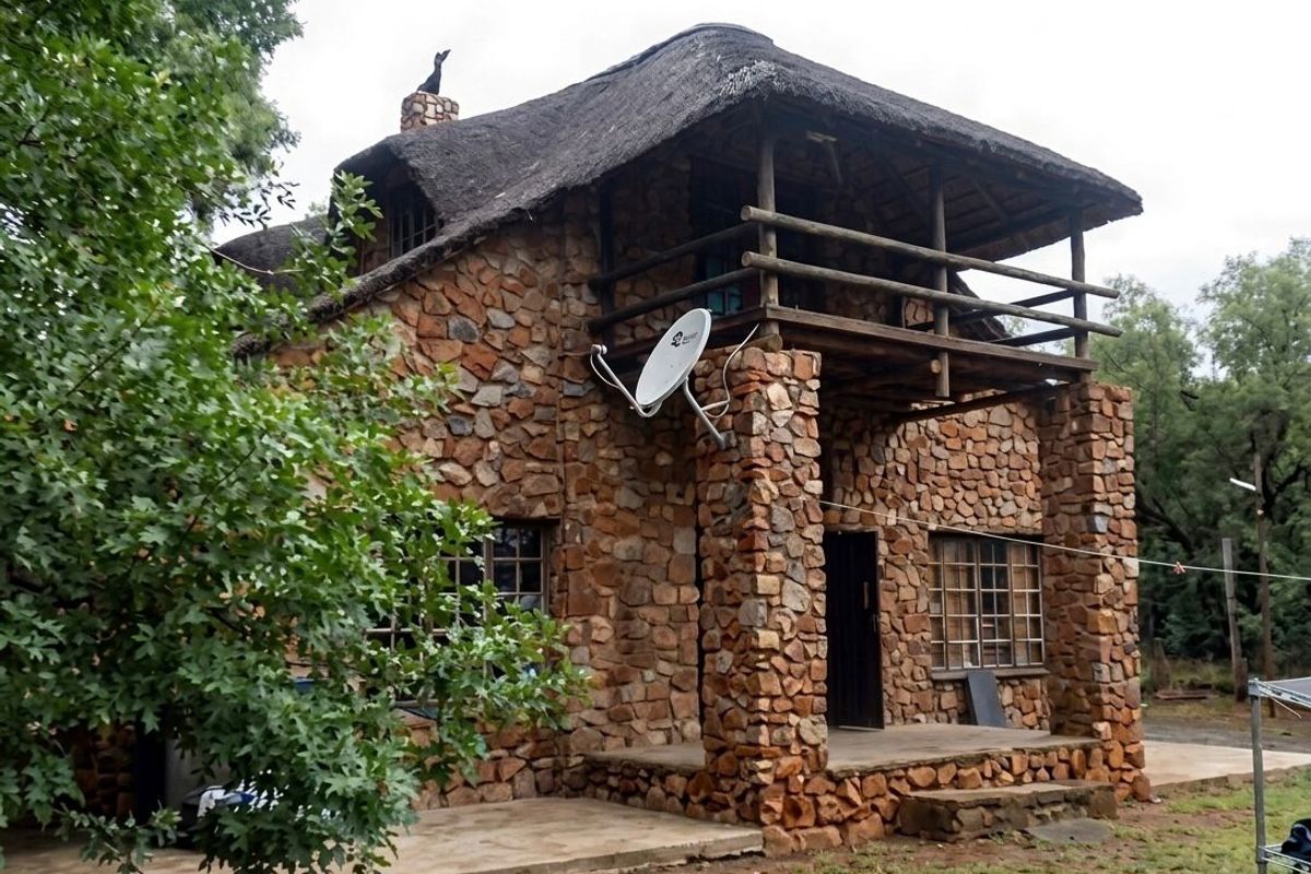 A rustic two-story stone farmhouse featuring a traditional thatched roof, a covered ground-floor patio, and an upper-level timber balcony.