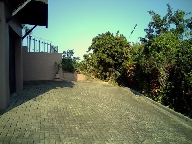 A paved driveway leading to a garage, featuring perimeter security fencing and established garden landscaping.