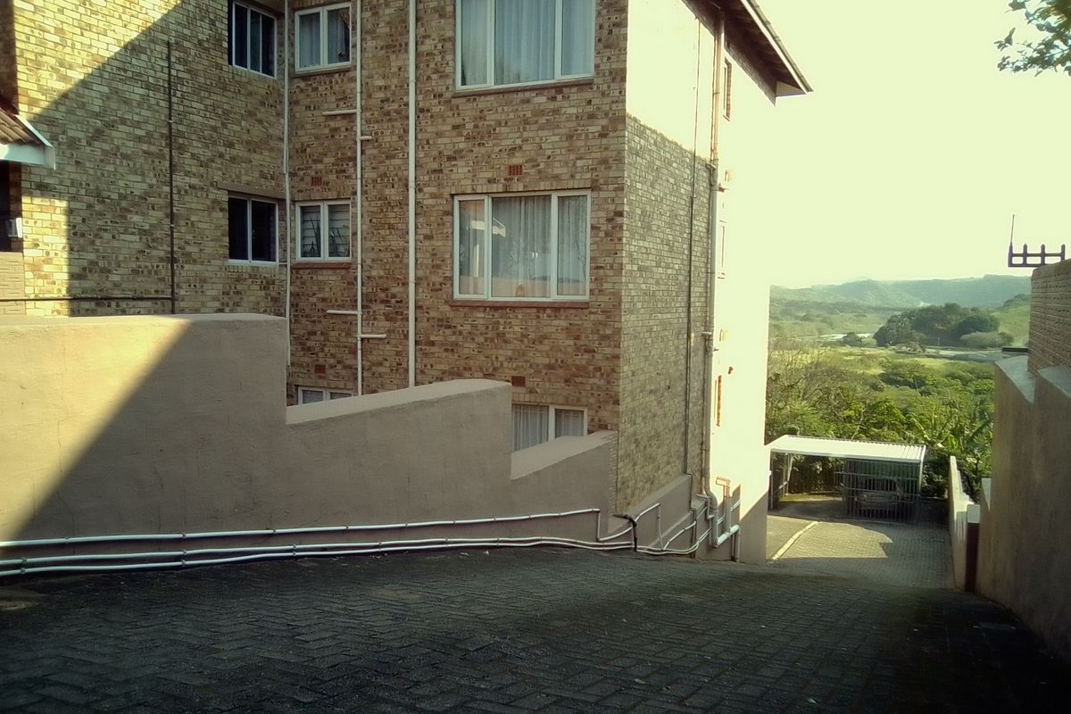 Rear view of a multi-story face brick apartment building featuring a paved driveway, retaining wall, and covered carport area.