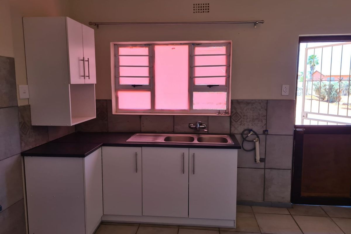 A functional kitchen area featuring white built-in cabinetry, a stainless steel sink, tiled splashbacks, and direct access to the exterior via a security gate.