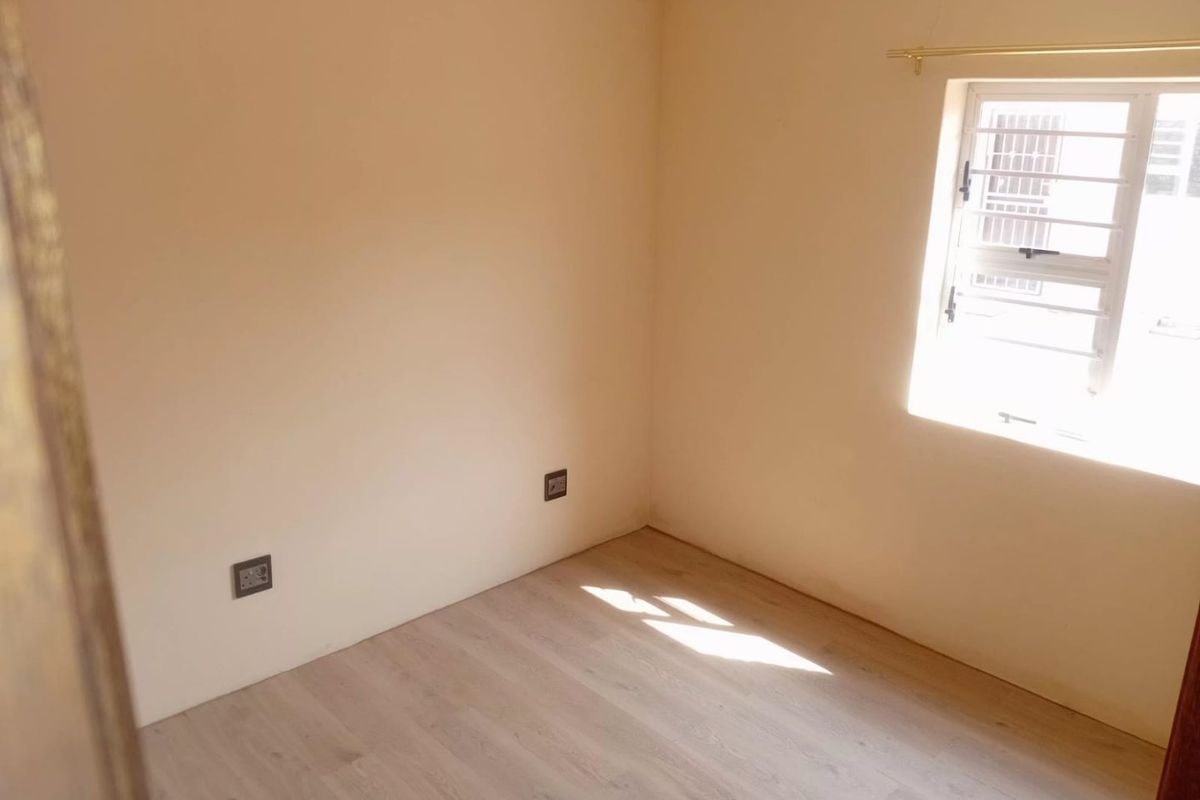 A bright, empty bedroom featuring light-toned laminate flooring, neutral walls, and a window fitted with security bars.