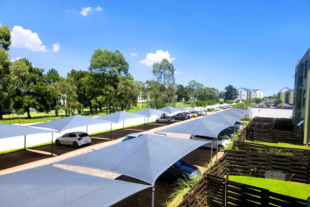 An elevated view of the apartment complex parking area featuring multiple shade cloth carports, paved driveway, and well-maintained timber fencing with lawn.