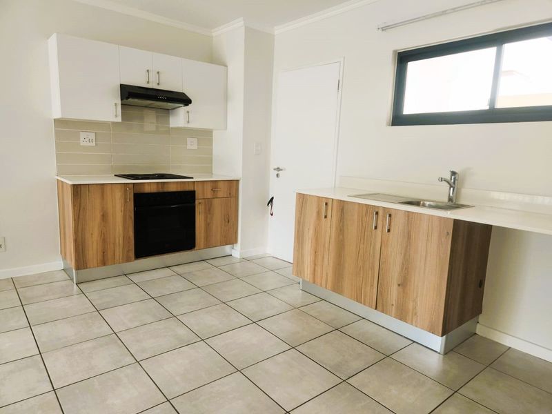 A clean, modern kitchen featuring wood-look cabinetry, white countertops, built-in oven and hob, and neutral tiled flooring in an open-plan layout.