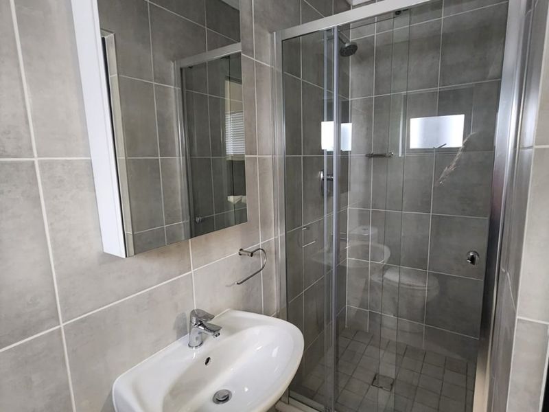 A clean, modern bathroom featuring a pedestal washbasin, a glass-enclosed shower, and neutral grey wall and floor tiling.